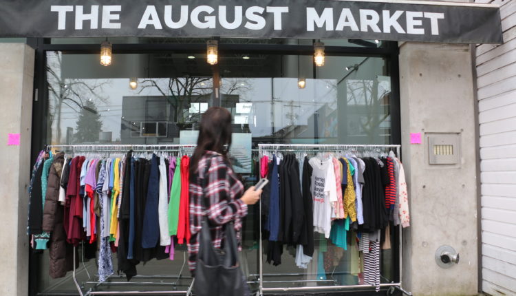 A woman holding a phone walks past racks of clothes on the sidewalk outside a retail shop.