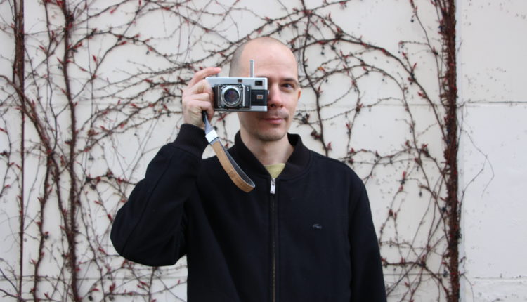 A man stands with an analogue film camera up to his face, his finger rests on the shutter button.