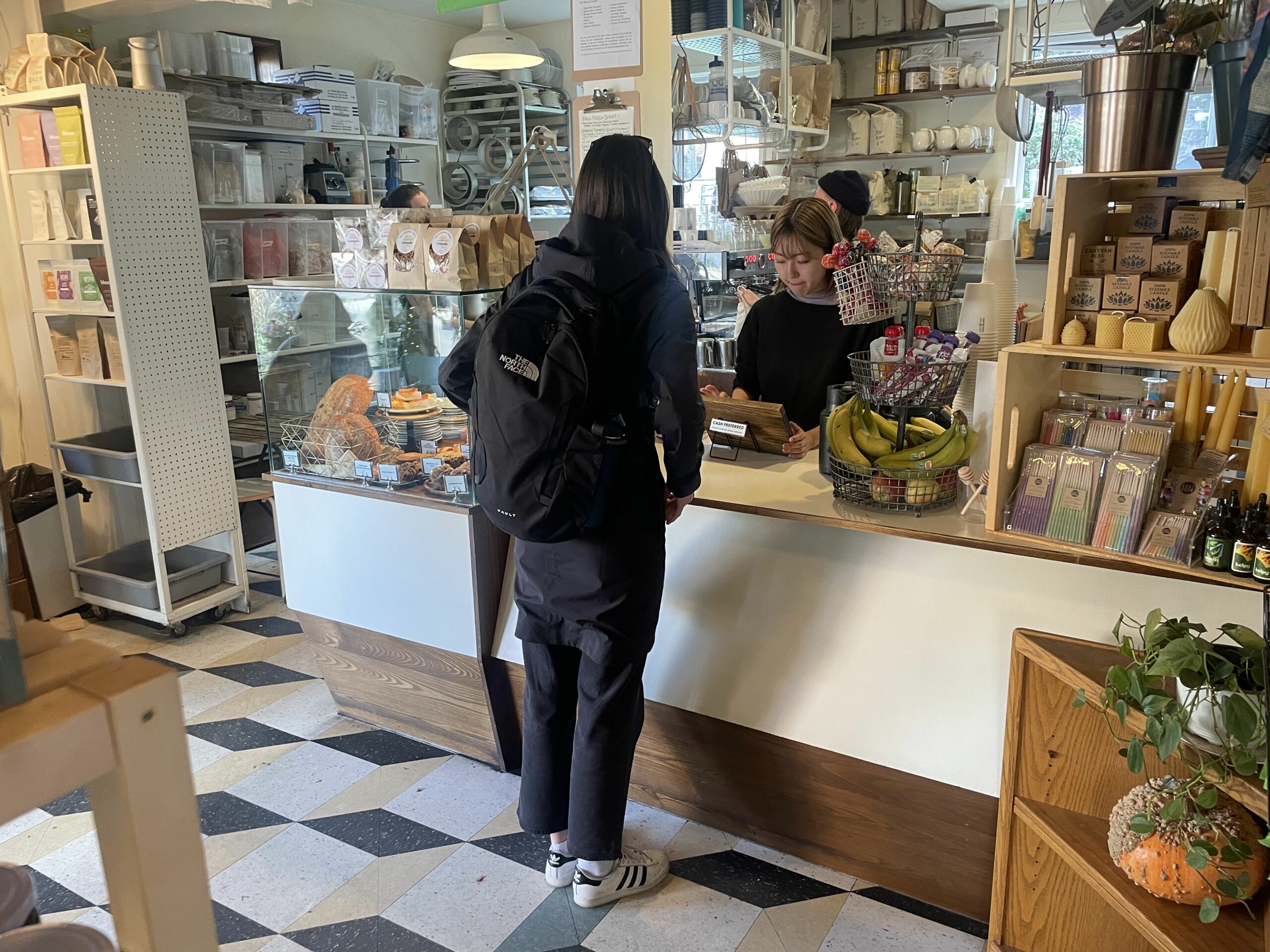 A person with a backpack is standing at a counter with a worker on the other side. The store has a black diamond, checkered floor and is filled with food and artisan items.