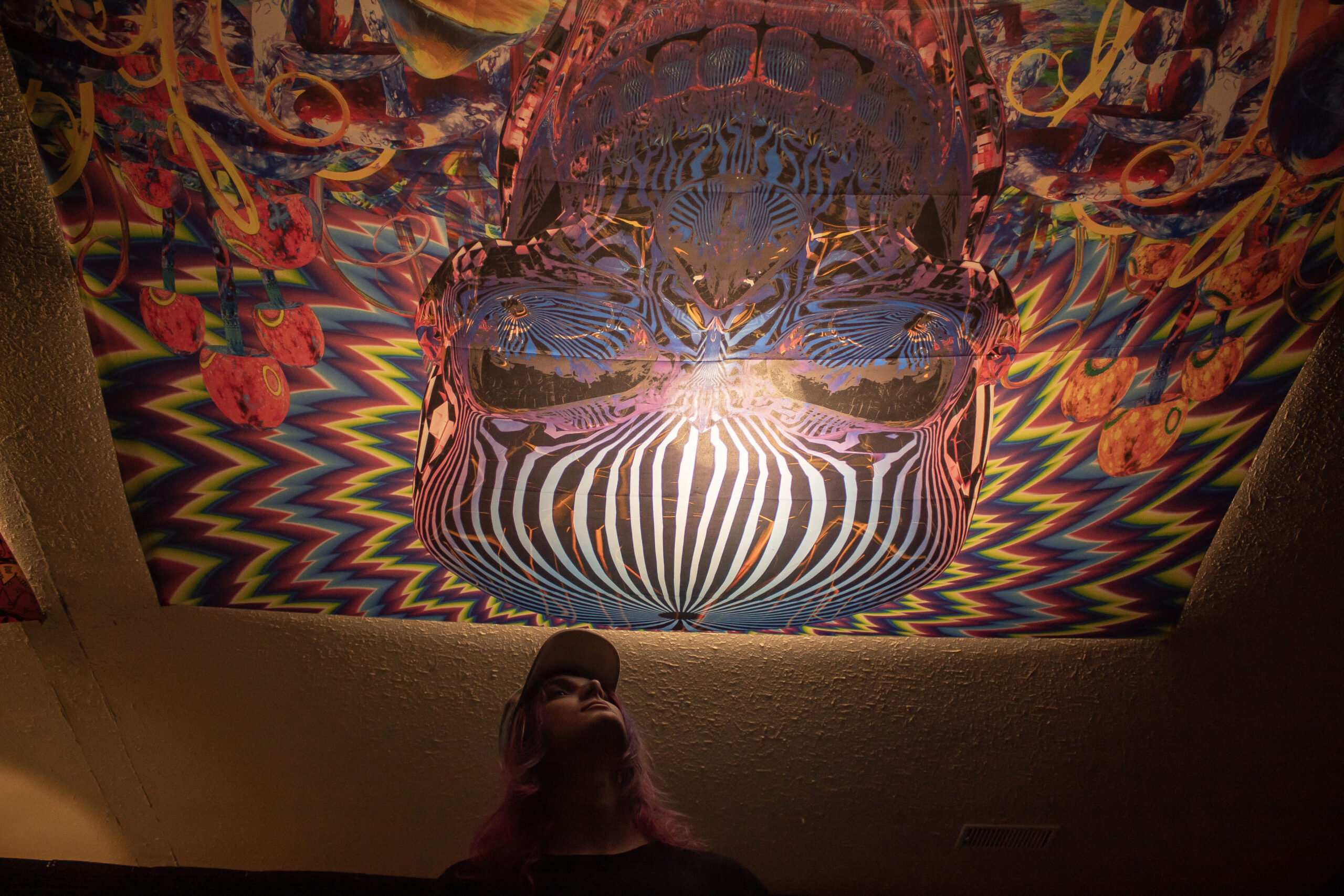 Angel Rivas looking up at a tapestry on his ceiling that depicts a large skull surrounded by colours