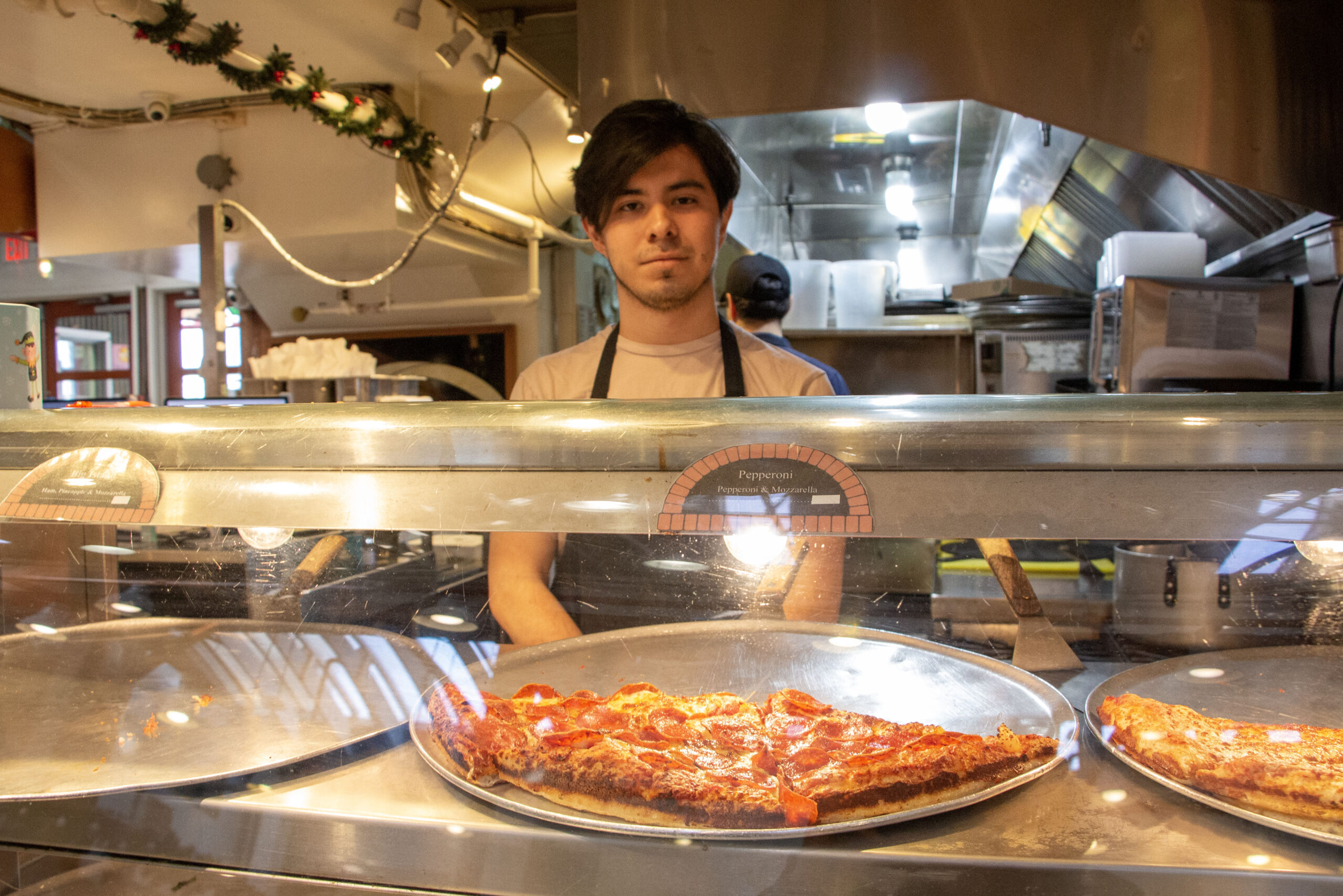 A man in an apron stands behind a display counter with a pizza inside of it.