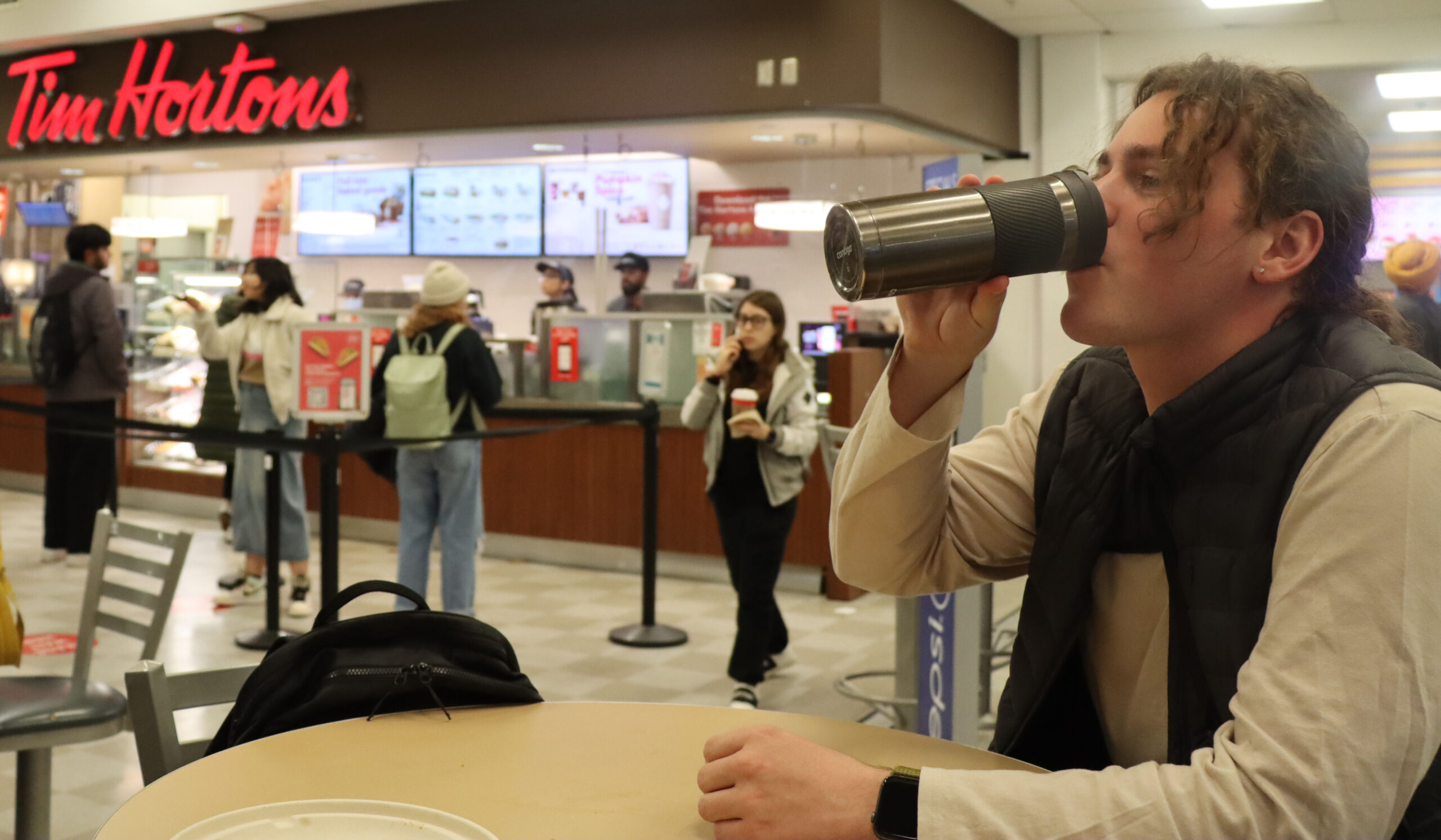A student sitting at a table drinking from a reusable mug. A Tim Hortons sign is seen in the background.
