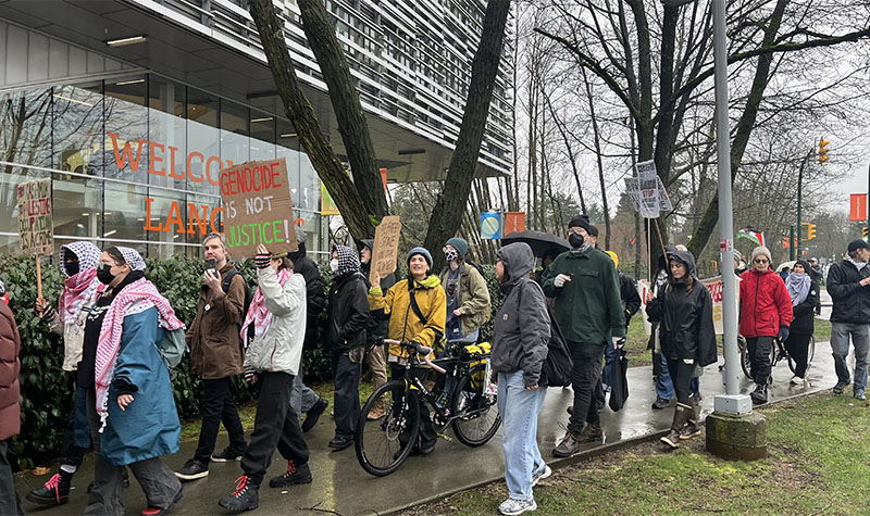 A crowd of protesters, holding signs, walk down a path by Langara's T building.